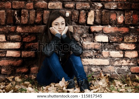 Lonely teenage girl sitting on the ground,a sad expression, leaning on an old brick wall.