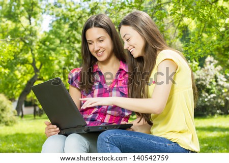 Two smiling female students using laptop in nature.