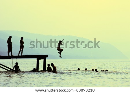 Silhouettes of kids who jump off dock on the lake at sunset.