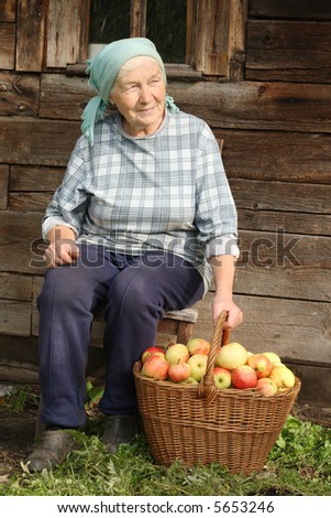 Senior countrywoman sitting against wooden wall with basketful of apples at her feet