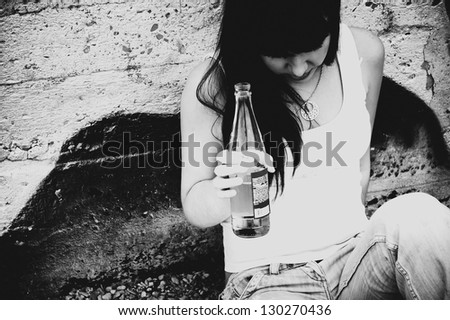 Drunk young girl with a bottle of beer in hand is sitting at the wall. Demonstration of youth social problems
