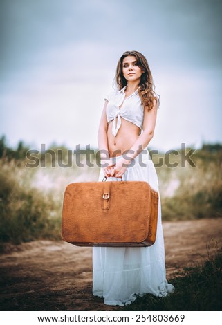 Beautiful young woman with suitcase in hands stands on a field road