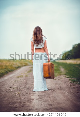 Young woman with suitcase in hand going away by a field road
