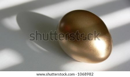 Golden egg on a white tabletop, with patterns of light and shadow laying across both.