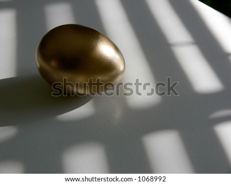 Golden egg on a white tabletop, with patterns of light and shadow laying across both.