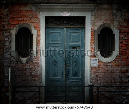 Crumbling weathered entrance to a building in Venice, Italy, with red brick wall, ornate barred windows and blue wooden door.