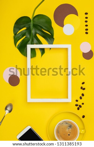 Creative flat lay with cup of coffee, paper circles, mobile phone, tropical plant and white frame for your text on yellow background