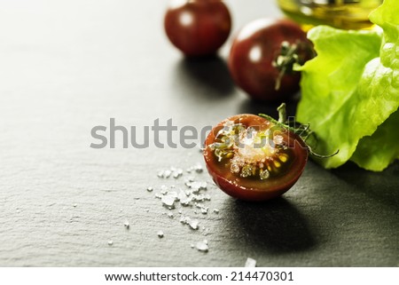 Fresh grape tomatoes with salad leaves and salt for use as cooking ingredients with a halved tomato in the foreground with copyspace