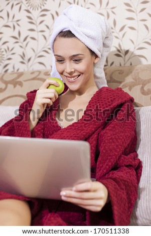 Beautiful girl in bathrobe eating apple and watching news on tablet pc