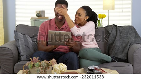 Young black woman covering boyfriend\'s eyes as he browses web on tablet