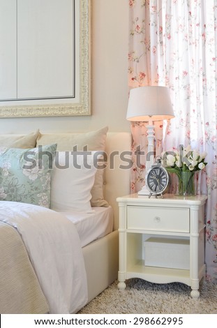 vintage bedroom interior with decorative table lamp, alarm clock and flower on white table