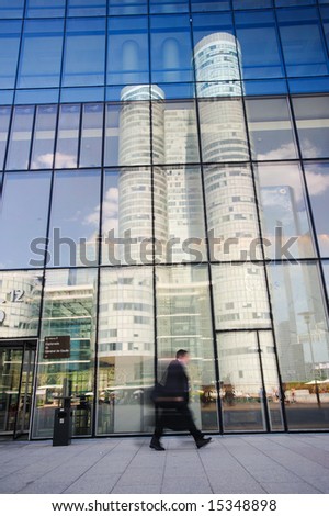 A businessman walking briskly near a office building reflecting another office building in its window glasses.