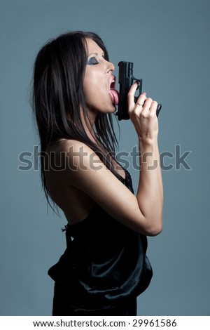 A portrait of a sexy female spy licking the tip of a gun, on turquoise studio background.