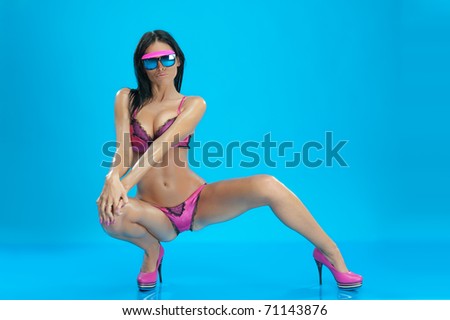 Sexy young woman with pink lingerie and sunglasses crouching in model pose, blue studio background.