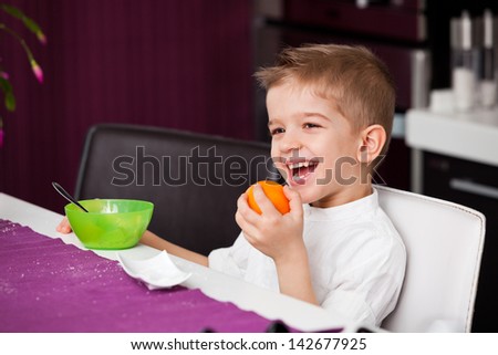 A five year old boy sitting at a kitchen table with an orange in his hand and a bowl in front of him. A five year old boy having breakfast