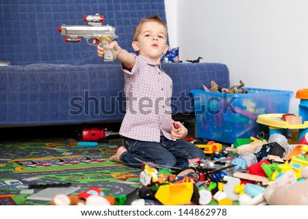 A five year old child playing in his room with a lot of toys around him. A five year old boy pointing at something/someone with his toy gun
