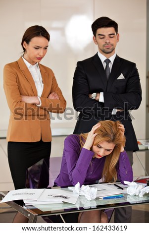 businesswoman holding her head in her hands, sitting at desk, with rumpled paper, coffee, phone, headache and angry colleagues watching her from the back - front view