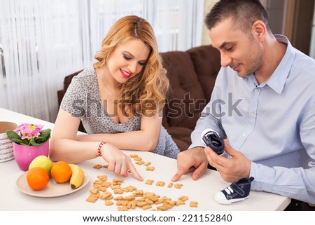 Pregnant young woman and her husband making their baby name using letters from biscuits, sitting at the kitchen table