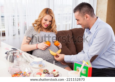 Picture of a young pregnant woman and her husband making a smile on her belly with a banana and two oranges, sitting at the kitchen table full of sweets