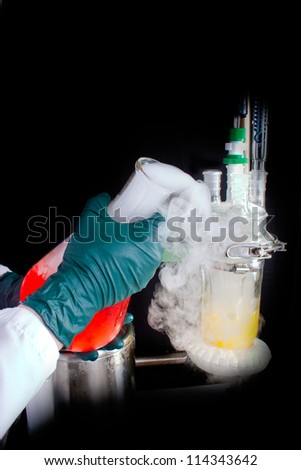 Close up of scientists hands, wearing plastic gloves, pouring a beaker filled with red chemical and vapors.