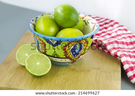 Limes in a colorful bowl on a wood cutting board with red and white dish cloth, white background