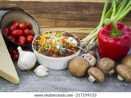 Bowl of Chili surrounded by ingredients of parmesan cheese, garlic cloves, tomatoes, scallions, mushrooms and red pepper with wood cutting board background