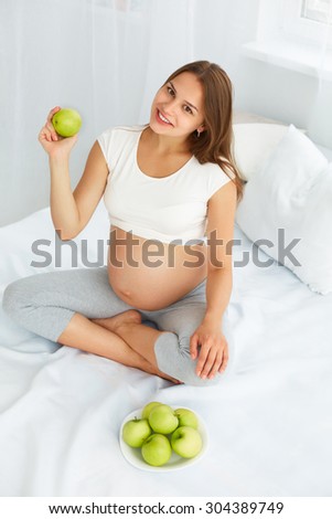 Pregnant Young Woman holding Apple while sitting on the Bed. Healthy Food and Diet Concept. Healthy Lifestyle.
