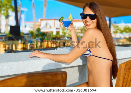 Summer party. Sexy young woman with long hair drinking cocktail at the beach bar in bikini and sunglasses.