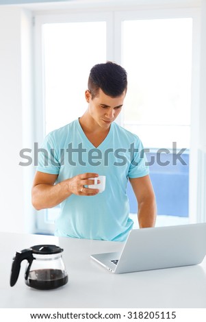 Handsome young man using a laptop pc in the kitchen while holding a cup of coffee in his hand