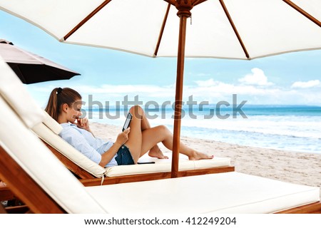 Beautiful Young Woman Reading E-book While Relaxing On Lounger Under Umbrella On Beach On Summer Holidays Vacations. Girl With Electronic Book Lying On Deck Chair Under Tent By Sea In Leisure Time