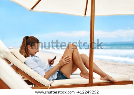 Portrait Of Beautiful Happy Smiling Woman Using Cell Phone And Dialing Number While Lying On Deck Chair On Beach By Sea. Girl On Summer Holidays Vacation. Mobile Communication, Connection Concept.
