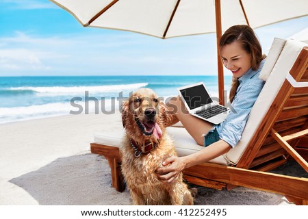 Beautiful Smiling Young Woman Playing With Pet While Using Laptop Computer Outdoors By Sea. Happy Girl Having Fun With Her Dog, Relaxing On Deck Chair At Beach On Summer Holiday Vacations.