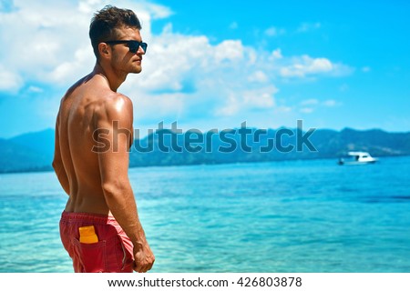 Summer Travel Vacation. Handsome Man With Sexy Body In Fashionable Sunglasses Sunbathing, Tanning At Sea Beach. Fitness Male Model With Sunscreen Lotion, Sun Block Skin Protection Cream In Pocket