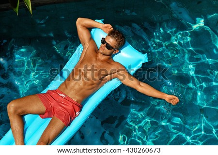 Man Summer Fashion. Beautiful Male With Sexy Body In Swimwear, Fashionable Sunglasses Tanning, Floating In Swimming Pool Water At Relax Spa Resort. Fitness Model With Skin Sun Tan Relaxing On Vacation
