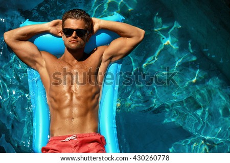Man Summer Fashion. Beautiful Male With Sexy Body In Swimwear, Fashionable Sunglasses Tanning, Floating In Swimming Pool Water At Relax Spa Resort. Fitness Model With Skin Sun Tan Relaxing On Vacation