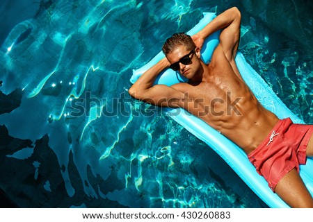 Summer Man Body Sun Skin Care. Beautiful Model With Sexy Body In Swimwear Tanning, Floating On Mattress In Swimming Pool Water. Fitness Male With Healthy Tan Relaxing At Relax Spa Resort. Summertime