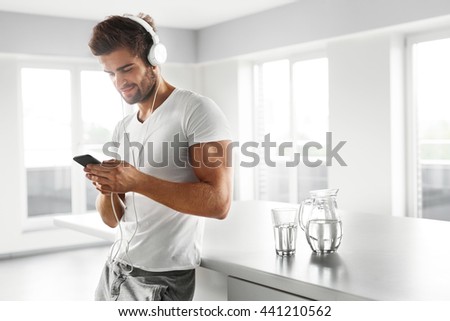 Man Listening To Music In Fashion Headphones Using Mobile Phone, Smartphone Indoors. Portrait Of Handsome Happy Relaxed Smiling Guy  Enjoying Music At Home. Entertainment, Communication Concept