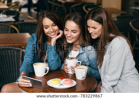 Young Female Friends Using Phone And Drinking Coffee In Cafe.. Beautiful Smiling Girls Enjoying Coffee And Biscuits, Using Mobile Phone While Sitting At Table In Coffee Shop. High Resolution.