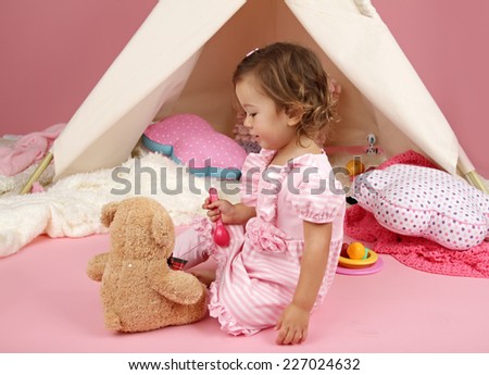 Happy toddler girl engaged in pretend play tea party with stuffed bear toy indoors at home with a teepee tent