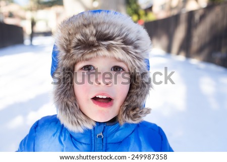 Child in a fuzzy fur snow hat and a winter jacket, face portrait, blowing snow at the camera, outside, outdoors