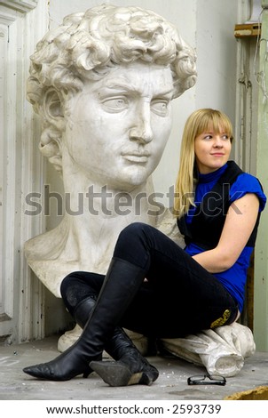 Blond girl in a studio next to clay statue of man's face looking away