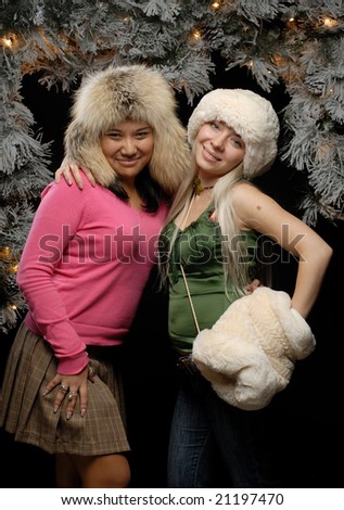 Two girls in fur hats against lit Christmas tree hugging smiling