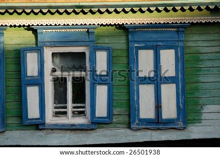A segment of an old green house with two windows with blue and white shutters one open the other closed
