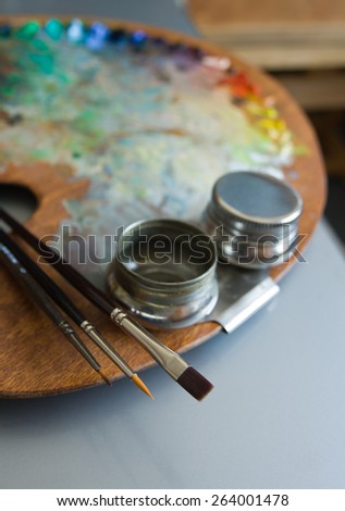 Paint palette. Wooden artist palette with three artists paintbrushes and steel twin dippers close-up on art studio interior background.