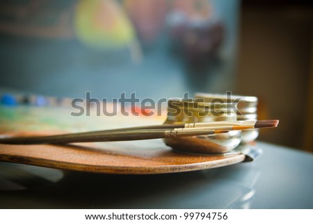 Painters palette. Wooden painter\'s palette with three artists paint brushes and steel twin dippers close-up on background of still-life painting in art studio interior.