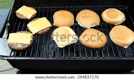 Hamburger patties with processed cheese slices cooking on an outdoor table top barbecue.  Buns toasting alongside