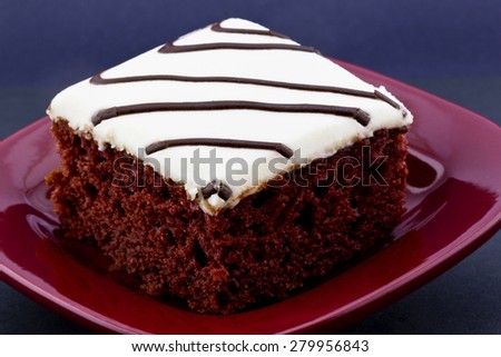 Red velvet cake with white icing and drizzles of chocolate placed on square, red plate on black background.