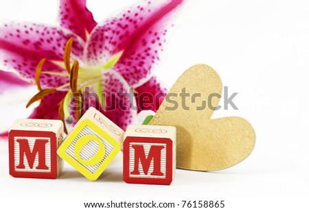 Gold heart and alphabet block letters spelling MOM are placed in front of a  single, lily blossom against a white background
