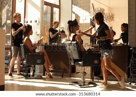 Picture of working day inside the beauty- sit on two chairs clients beautiful young girls. Hairdresser makes hair styling or hair cut, make-up artist doing make-up in a beauty salon- stock photo.
