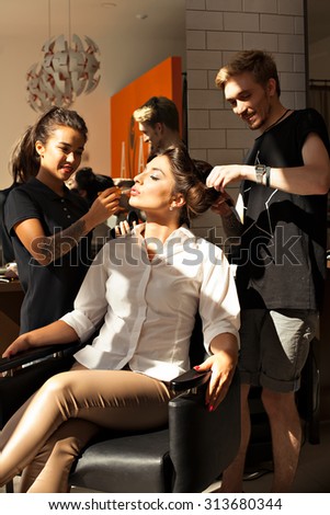 The image of a beautiful young brunette girl model sitting in a chair in a beauty salon with a hairdresser and makeup artist. Stylist- hairdresser and stylist, makeup artist working - STOCK PHOTO